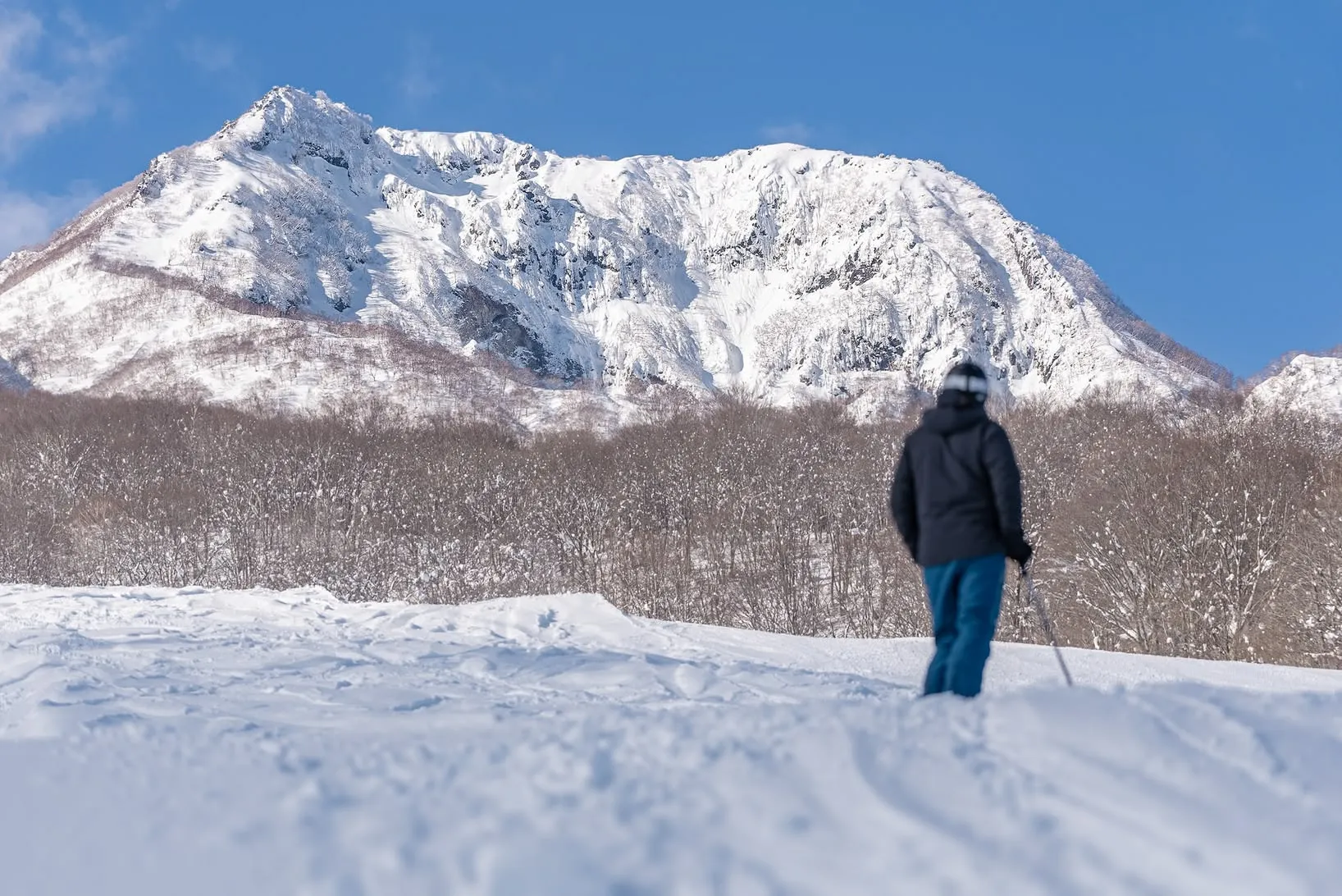 Skier standing on a snowy slope looking across the valley towards Mount Myoko under a clear blue sky